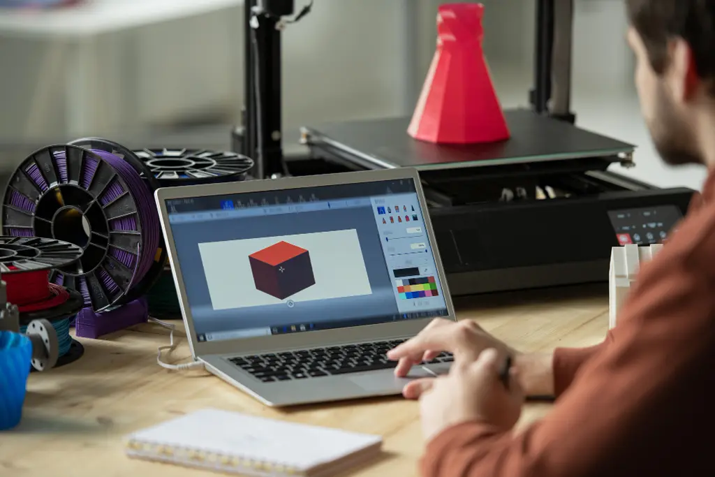 Conception et numérisation 3D – une personne travaille sur un ordinateur portable avec un modèle 3D d'un cube, à côté d'une imprimante 3D, symbolisant des services complets d'impression 3D.